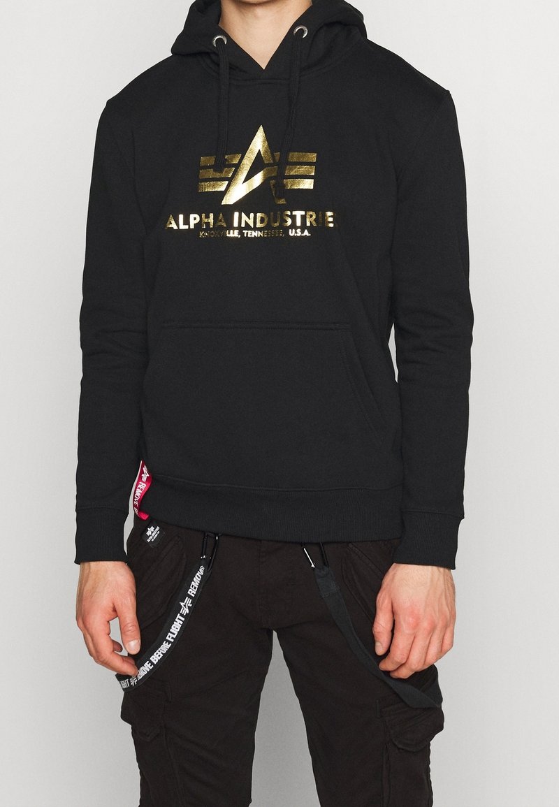 Felpa nera con cappuccio con il logo dorato "ALPHA INDUSTRIE" sul petto. Presenta una tasca a marsupio e un cappuccio con stringa, con un'etichetta di marca sul lato.