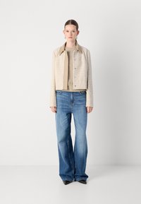 Beige strukturierte Jacke mit kragenloser Ausschnitt, über einem hellbraunen gestrickten Oberteil. Lockere blaue Jeans mit weitem Bein und schwarzen spitzen Schuhen.