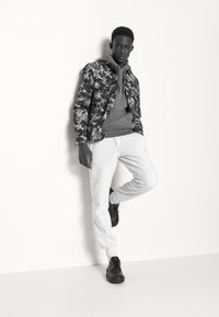 Casaco preto com padrão floral sobre um hoodie cinza, combinado com calças de moletom brancas e sapatos pretos com atacadores; modelo inclina-se contra uma parede.