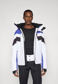 Biela prešívaná bunda s modrými a čiernymi akcentmi, predný zips, veľká kapucňa a manžety s otvorom na palec. Nosená cez čiernu dlhú rukávovú košeľu.