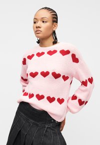 Νεαρό άτομο με κοτσίδες που φοράει ροζ πλεκτό πουλόβερ με κόκκινα σχέδια καρδιών και μαύρη πλισέ φούστα, κοιτάζοντας προς το πλάι.