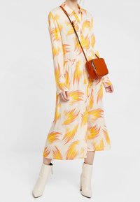 Vestito a maniche lunghe color crema con motivi di foglie arancioni e gialle, vita aderente, abbinato a una piccola borsa arancione a tracolla e stivaletti bianchi.