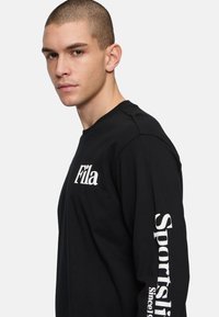 Camiseta negra de manga larga hecha de algodón. Presenta el logo de "Fila" en el pecho y el texto "Sportslip Since" a lo largo de la manga izquierda.