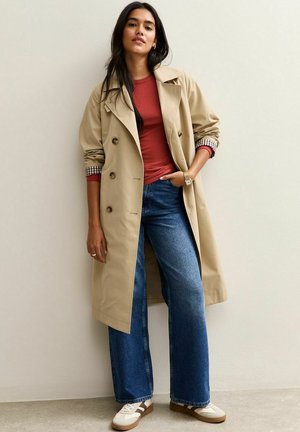 Vrouw staat tegen een muur gekleed in een beige trenchcoat, een rustkleurig lange mouwen shirt, wijdvallende blauwe jeans en witte sneakers met bruine accenten.