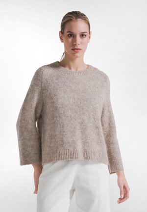 Kvinde med lysebrunt hår sat op i en hestehale iført en løs beige striktrøje og hvide bukser, stående foran en ensfarvet hvid baggrund.