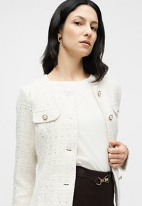 Veste en tweed blanc avec tissu texturé, poches avant et boutons ressemblant à des perles. Portée sur un haut clair et un pantalon tailleur foncé.