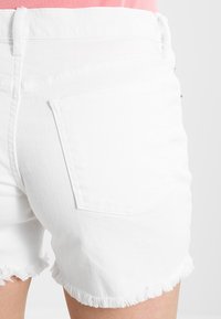 Shorts de mezclilla blanca con dobladillo deshilachado, que cuentan con bolsillos traseros y una cinturilla resistente. La tela parece gruesa y texturizada.