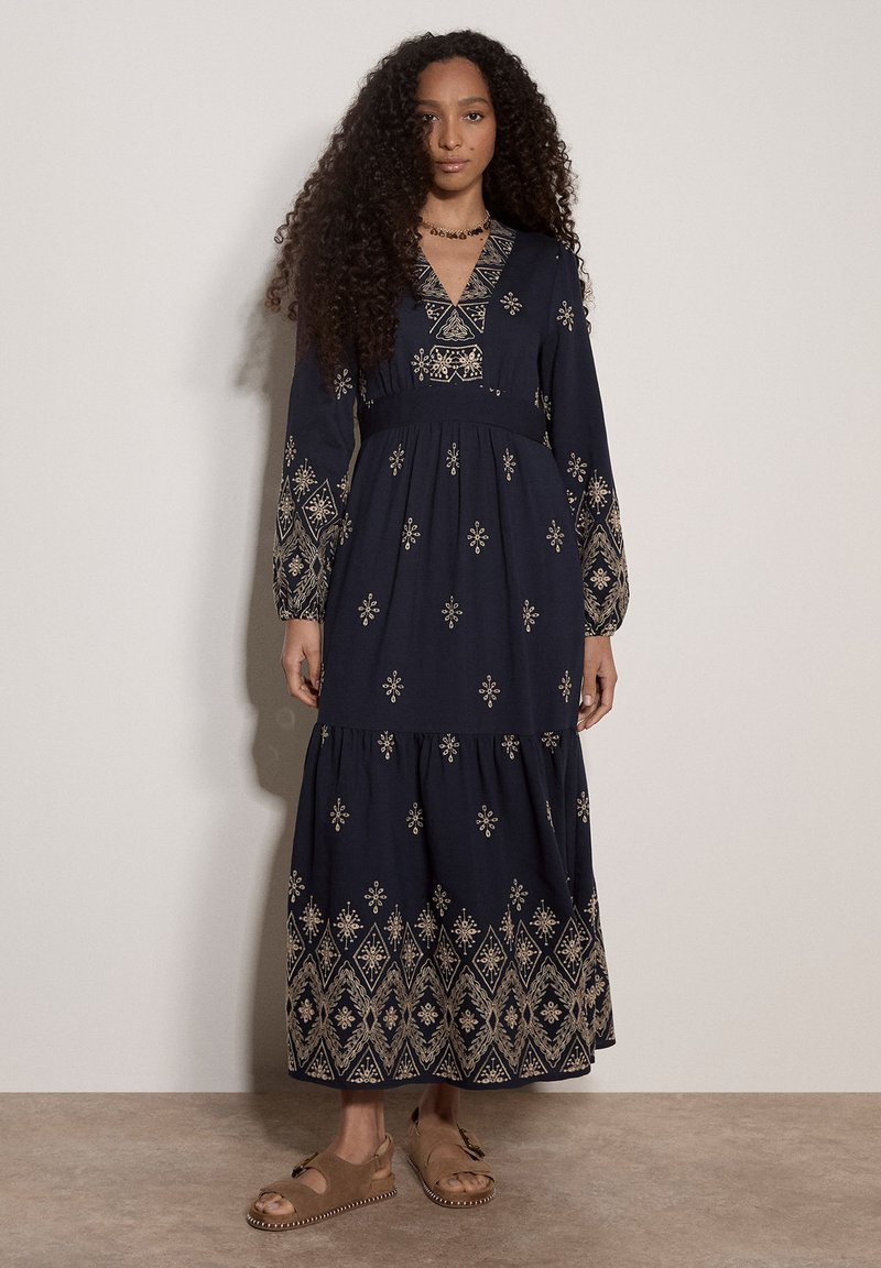 Femme aux longs cheveux bouclés portant une robe longue bleu marine avec une broderie géométrique beige et des sandales couleur caramel, debout contre un mur uni.