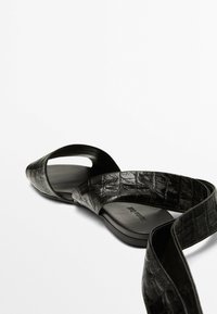 Schwarze flache Sandalen mit strukturiertem Kunst-Krokodilfinish, spitzer Zehenform und breitem Riemen. Die Sohle ist glatt und hat ein minimalistisches Design.