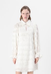 Robe blanche avec un motif zigzag texturé, fermeture à boutons et un col. Manches longues avec des poignets à volants.
