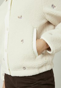 Veste en polaire de coloris écru avec broderie florale. Comprend des poignets côtelés et une fermeture à boutons. Inclut des poches latérales pour le rangement.