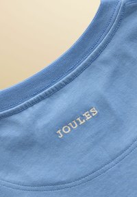 Gros plan sur l'encolure en tissu bleu clair avec le nom de la marque "JOULES" brodé en blanc, centré sous la couture du col.