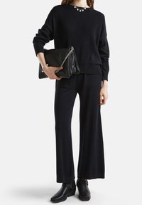 Czarny sweter dziany z prążkowaną fakturą, zestawiony z szerokimi czarnymi spodniami. Trzyma czarną skórzaną torebkę na ramię z łańcuszkiem i nosi botki za kostkę.