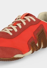 Chaussure de sport rouge et beige avec des lacets blancs, des détails en suède et une semelle brune texturée avec un design angulaire sur le côté.