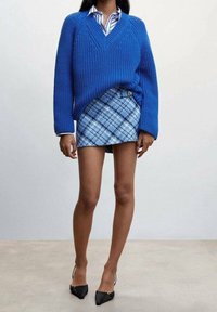 Femme portant un pull en tricot bleu à col en V sur une chemise rayée, assorti à une mini-jupe à carreaux bleue et des talons pointus noirs.
