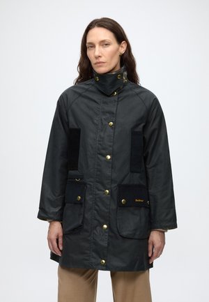 Femme aux longs cheveux foncés portant une veste noire boutonnée avec un col haut et de grandes poches avant, debout devant un fond blanc uni.
