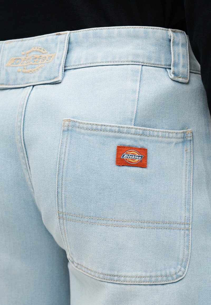 Hellblaue Denim-Jeans mit Gesäßtasche, orangefarbenem Dickies-Label und passender Denim-Gürtelschlaufe, getragen mit schwarzem Oberteil.