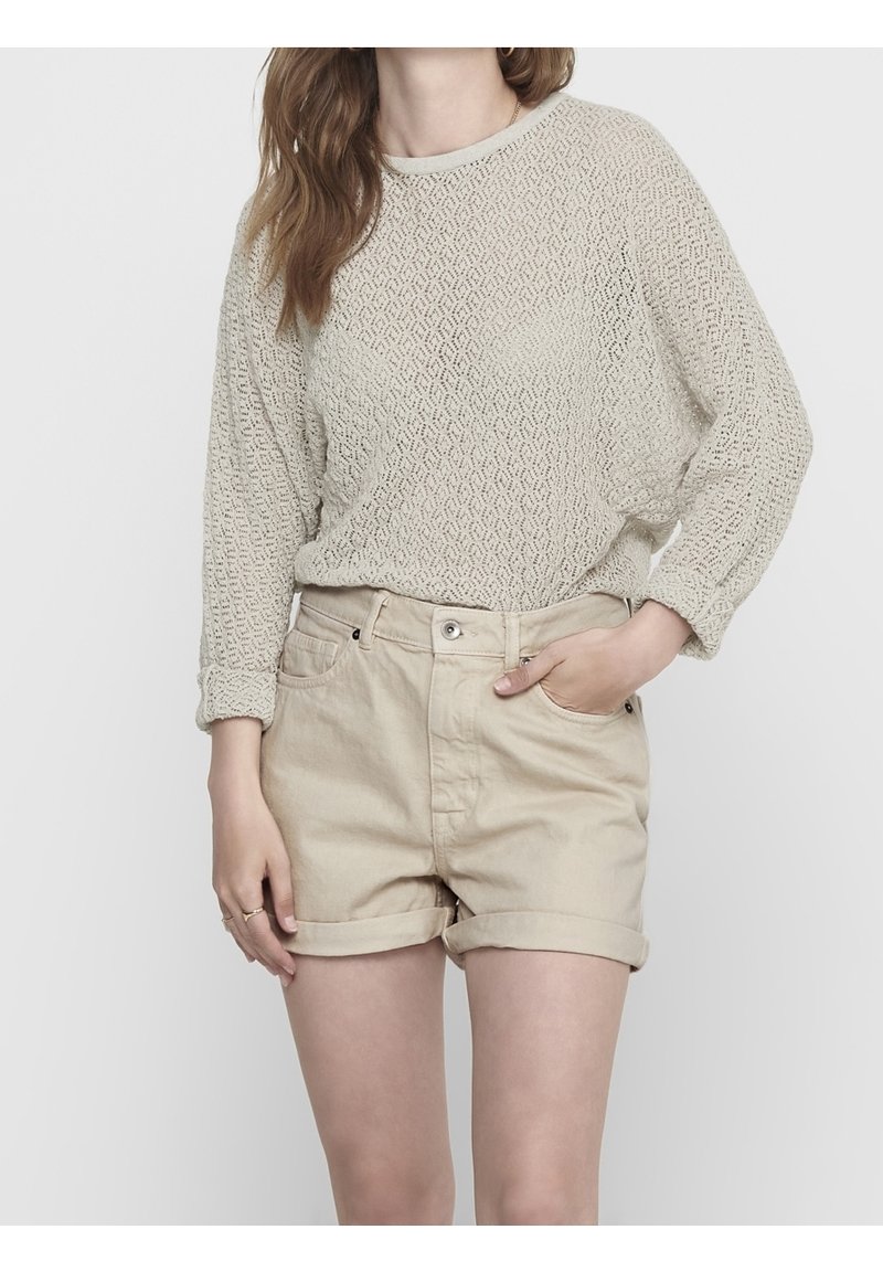 Femme portant un haut à manches longues beige texturé et un short beige roulé, une main dans une poche et l'autre reposant sur le côté.