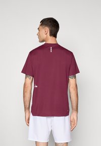 Maglietta sportiva a maniche corte color bordeaux con colletto tondo, texture liscia e dettagli a pois bianchi sulla schiena e sul lato.