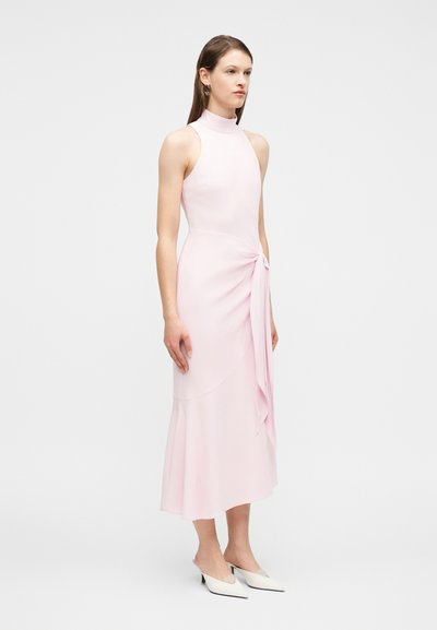Γυναίκα που στέκεται φορώντας αμάνικο, απαλά ροζ, μίντι φόρεμα με ψηλό λαιμό και λεπτομέρεια κόμβου στο πλάι, συνδυασμένο με λευκά παπούτσια με χαμηλό τακούνι.