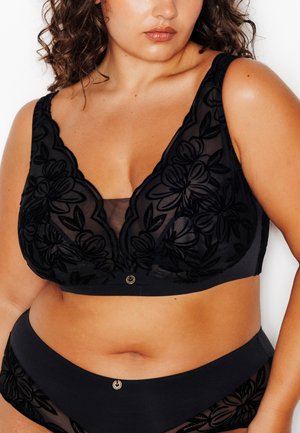 Donna formosa che indossa un reggiseno nero in pizzo floreale e mutandine a vita alta abbinate, ritagliata dalle spalle ai fianchi su sfondo bianco.