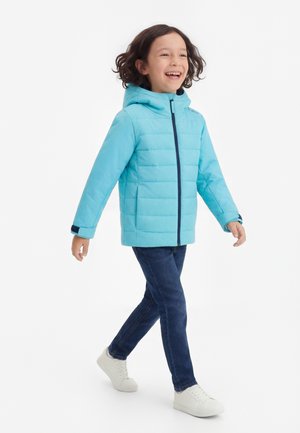 Dziecko idące z radosnym wyrazem twarzy, ubrane w jasnoniebieską kurtkę z kapturem, ciemnoniebieskie dżinsy i białe trampki na białym tle.