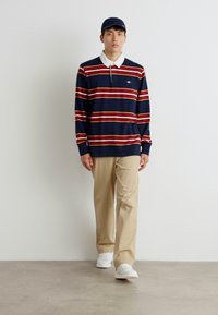 Marine stripete langermede polo-skjorte med rød- og hvitstripete, hvit krage, beige bukser og hvite sneakers. Modell i en minimal setting.