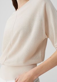 Hellbeige gestrickter Pullover mit kurzen Ärmeln, geripptem Saum und subtiler Textur. Verfügt über eine zentrale Naht an der Vorderseite.
