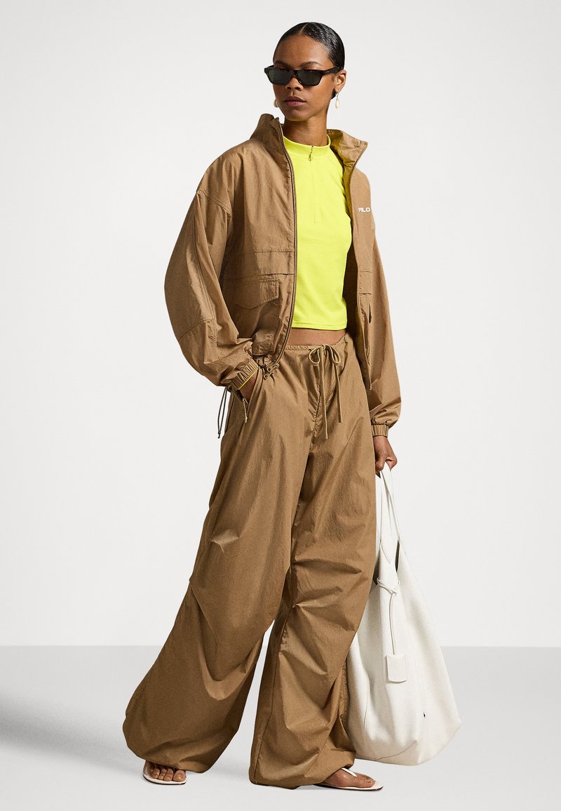 Frau trägt übergroße braune Jacke und Hose, neon-gelbes Oberteil, schwarze Sonnenbrille, hält eine große weiße Einkaufstasche, barfuß auf weißem Boden.