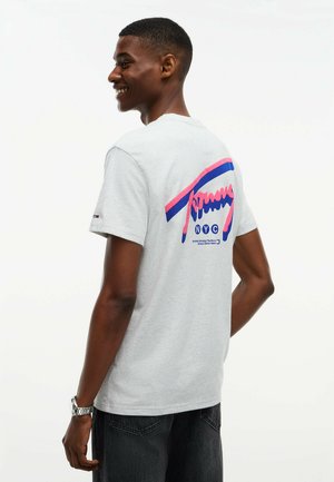Junger Mann lächelt, trägt ein hellgraues T-Shirt mit buntem Grafikmotiv und dem Text "NYC" auf dem Rücken sowie dunkle Jeans.