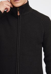 Schwarzer gestrickter Reißverschluss-Pullover mit hohem Kragen, der über eine gerippte Textur und ein kontrastierendes glattes Reißverschlussdetail verfügt.