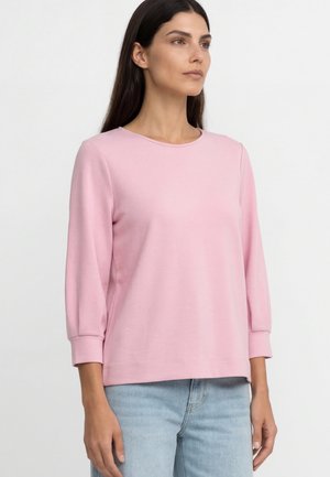 Kobieta z długimi ciemnymi włosami, ubrana w jasnoróżową bluzkę z długim rękawem i jasnoniebieskie dżinsy, stojąca na jednolitym białym tle.