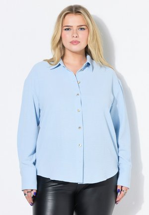 Blondynka w jasnoniebieskiej koszuli zapinanej na guziki i czarnych skórzanych spodniach, stojąca na białym tle.