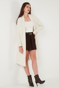 Crème ribgebreide cardigan met open voorkant, gedragen over een witte top en zwarte rok, geaccessoireerd met een zwarte riem en bruine enkelhakken.