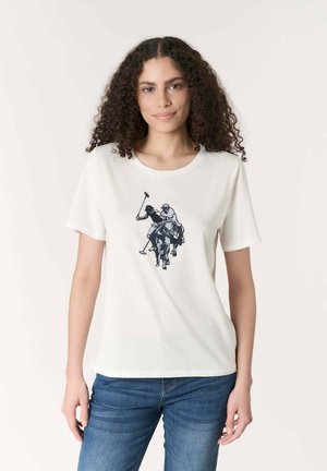 Donna con capelli ricci che indossa una maglietta bianca con una grafica nera raffigurante due giocatori di polo a cavallo, abbinata a jeans blu su sfondo semplice.