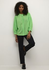 Blouse verte, légère, à pois avec un devant boutonné et des manches bouffantes, associée à un pantalon noir à jambe droite et des chaussures noires.
