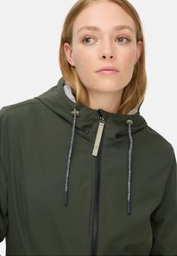 Grøn hættejakke med lynlås foran, grå inderside og teksturerede snore. Glat stof med et tætsiddende design.