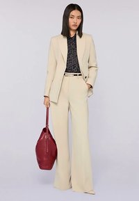 Beige op maat gemaakt broekpak met een aansluitend jasje en wijd uitlopende broekspijpen. Zwarte blouse met patroon eronder, met een bordeauxrode handtas in de hand.