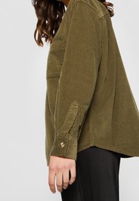 Femme portant une chemise en velours côtelé vert olive à manches longues avec des poignets boutonnés et un pantalon noir à côtes, debout devant un fond uni.