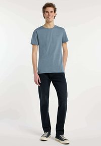 T-shirt bleu à manches courtes avec un col rond et un petit logo, associé à un jean foncé et des baskets en toile. Texture lisse, design décontracté.