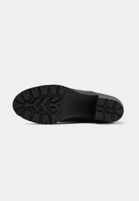 Suela de zapato negra con patrones de agarre profundos, que presentan formas en zigzag y rectangulares sobre un fondo blanco liso.