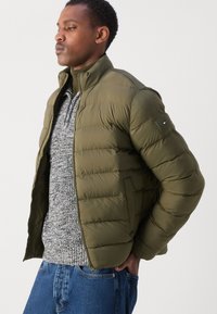 Olivegrøn pufferjakke med høj krave, der har en lynlåslukning foran og en tekstureret grå sweater under. Skræddersyet pasform, glat finish.
