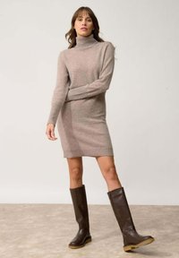 Abito a collo alto in maglia beige chiaro, di lunghezza a mezza coscia, abbinato a stivali marroni scuri sopra il ginocchio, con suola piatta e pelle testurizzata.