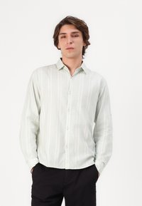 Chemise boutonnée vert menthe clair avec des rayures blanches verticales, manches longues et une texture de tissu douce. Associée à un pantalon sombre.