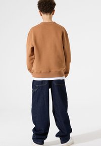 Felpa in pile marrone con colletto a girocollo, cuciture laterali e polsini a costine. Abbinata a jeans in denim blu scuro a vestibilità ampia.