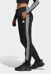 Čierne športové nohavice s bielymi pruhmi po stranách, elastický pás a vrecká, v kombinácii s čiernymi bežeckými topánkami.