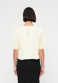 Femme aux cheveux courts et foncés portant un chemisier blanc à manches courtes bouffantes et broderie florale, associé à une jupe noire côtelée, vue de dos.