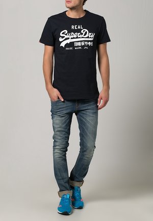 Camiseta azul marino de manga corta con texto de marca blanco, combinada con jeans de mezclilla azul claro y zapatillas azules con detalles en rosa. Ajuste casual.