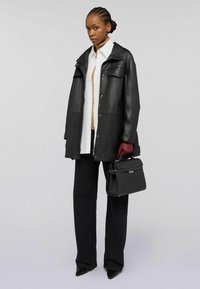 Donna che indossa una giacca di pelle nera, una camicia bianca, pantaloni neri, guanti rossi e tiene in mano una borsa nera con texture su uno sfondo grigio uniforme.