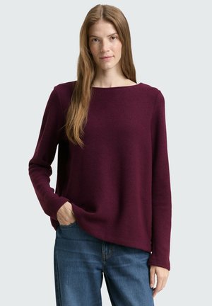 Bordeaux trui met een brede halslijn, lange mouwen en een gemêleerd breisel. Gepaard met blauwe denim jeans, model staande tegen een neutrale achtergrond.